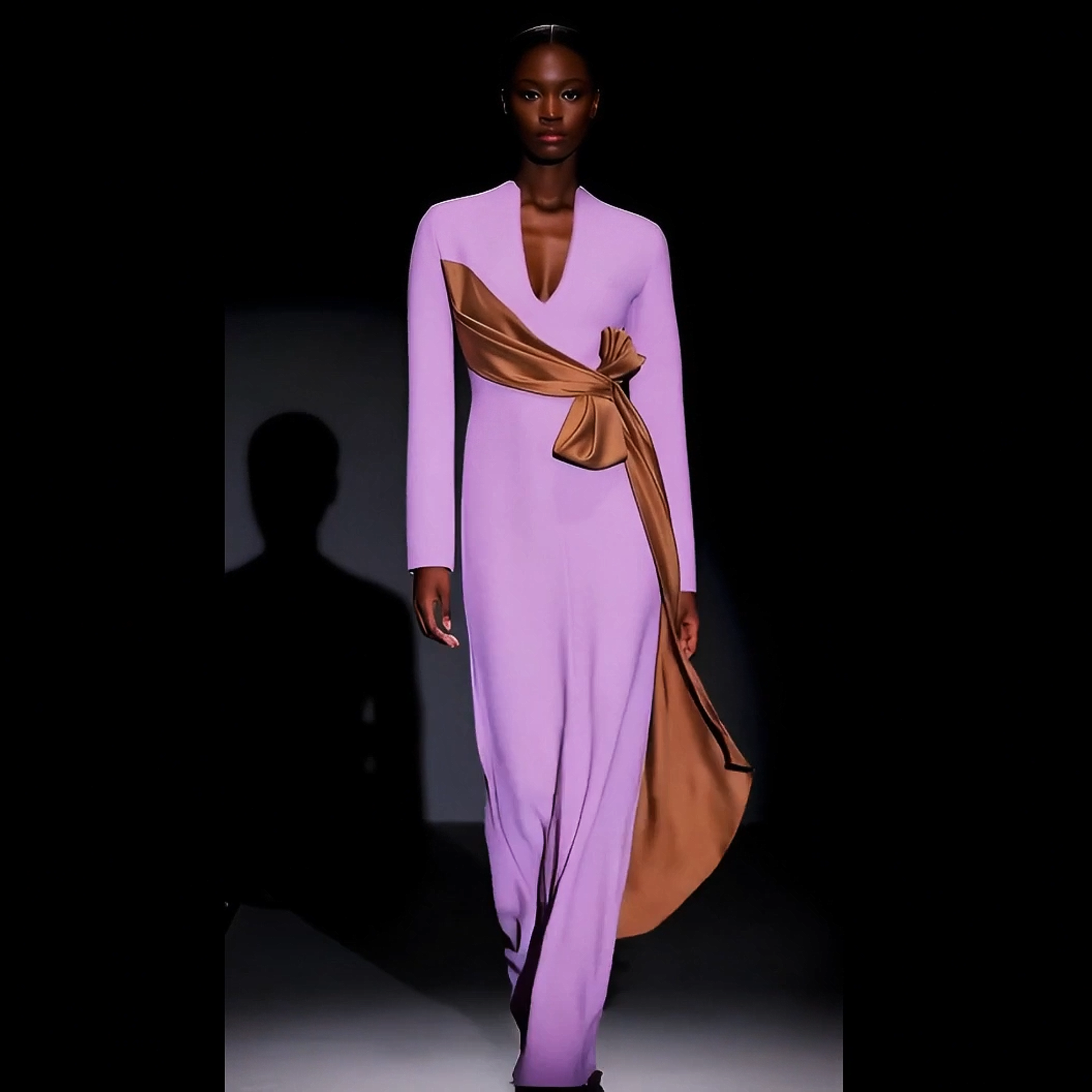 Model wearing a long, light purple dress with a brown sash on a dark background